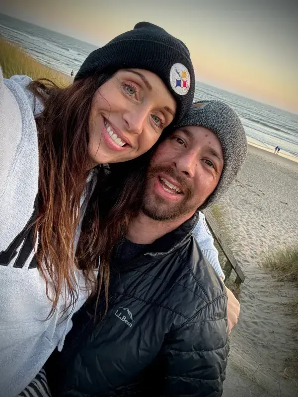 Couple smiles on beach, one wearing a Steelers beanie, other in a gray hat, embracing. Sunset background.