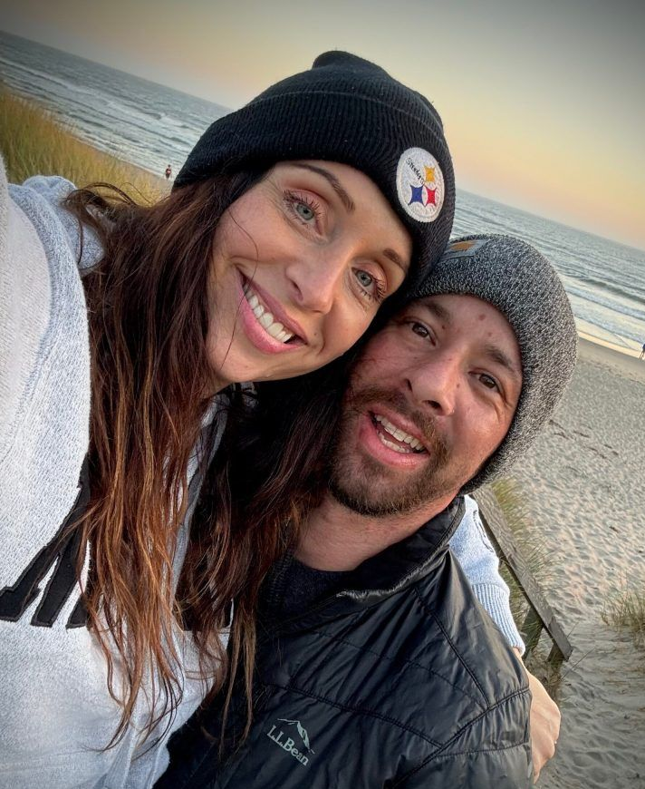 Couple smiling together at a beach, woman in Steelers beanie, man in grey hat.