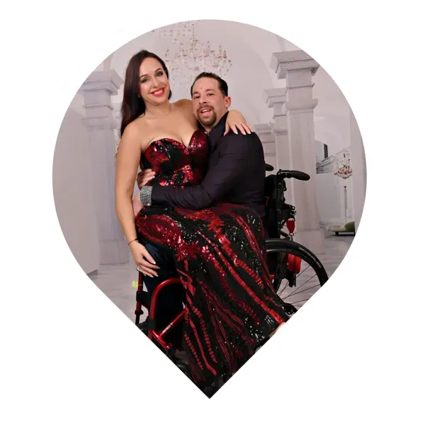 Woman in red sequined dress embraces man in wheelchair, formal setting.