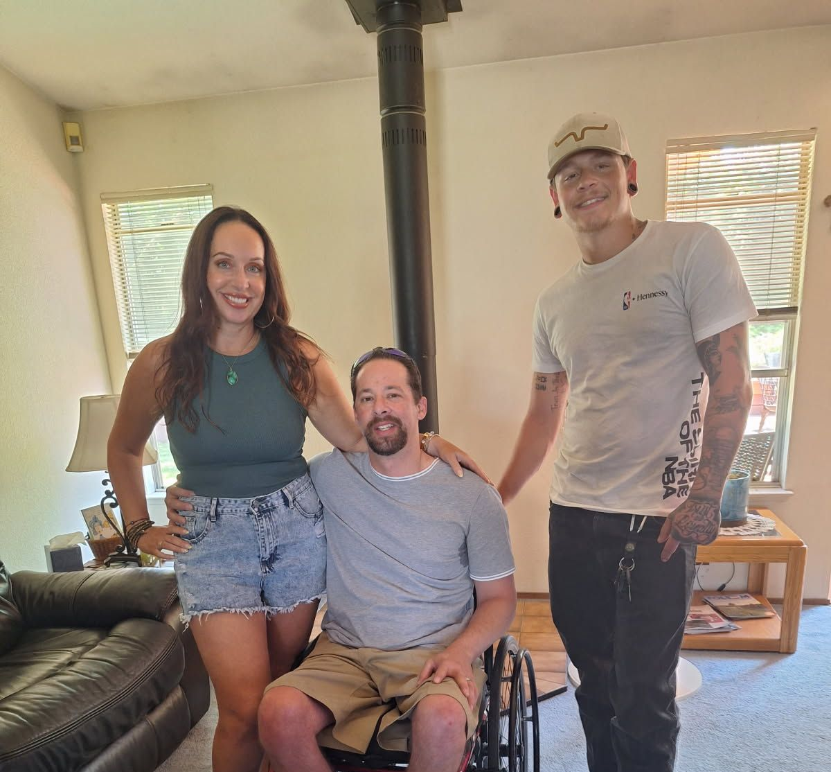 Three people posing indoors; a woman with arm around a man in a wheelchair, another man stands beside them.