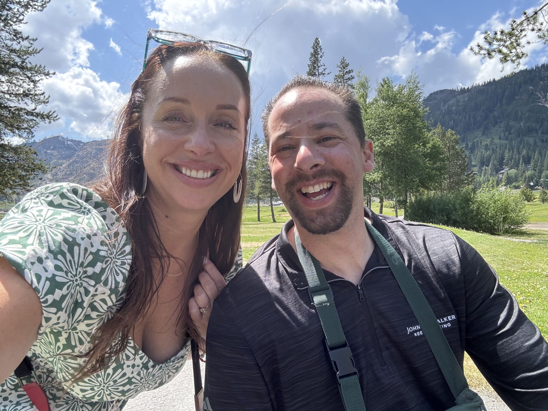 Smiling couple taking a selfie outdoors with mountains in the background. Sunny day.