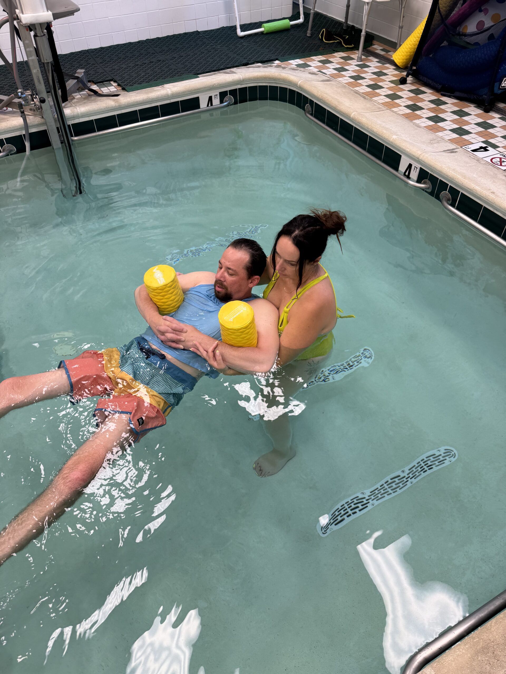 Man in pool with flotation devices, supported by a woman. They are in a therapeutic aquatic setting.