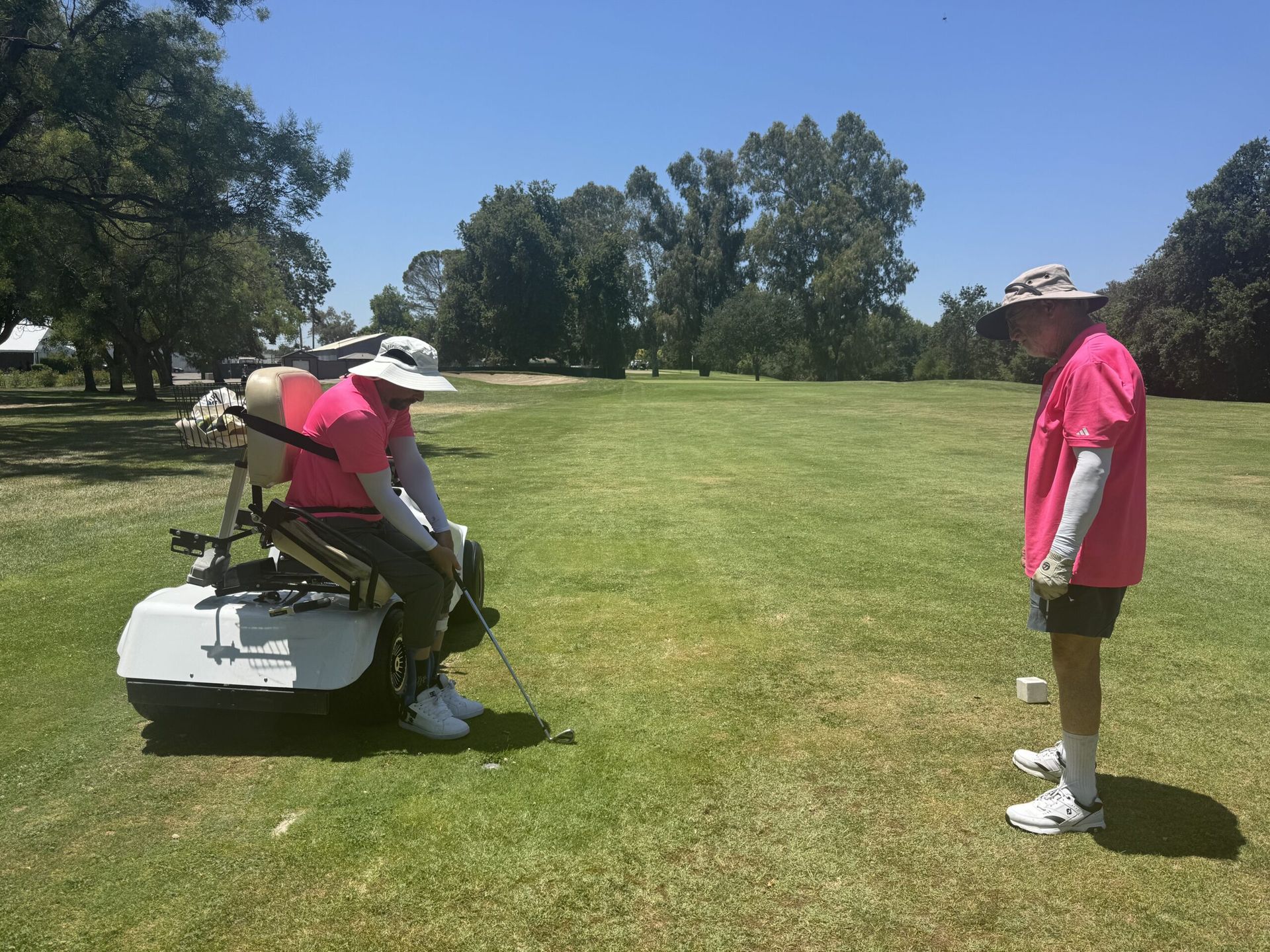Two people golfing: one in a specialized chair, the other standing. Both wear pink shirts and hats on a green course.
