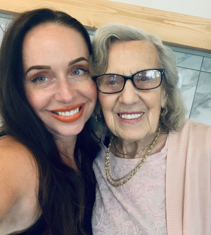 Woman and older woman smiling, taking a selfie. Both have light skin, the older woman wears glasses and a necklace.