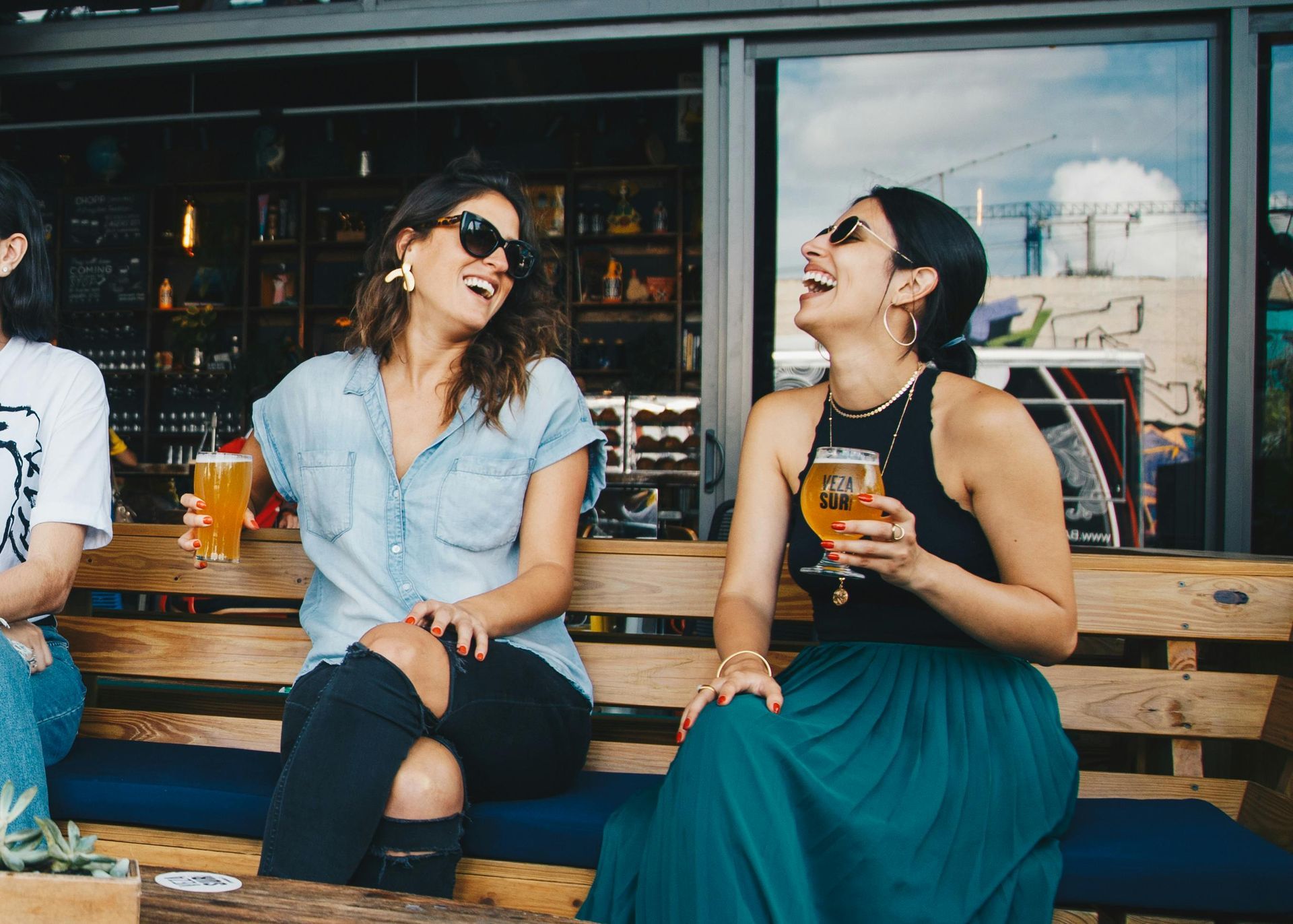 Two women laughing, seated outdoors, each holding a drink. One in denim, the other in a teal skirt.