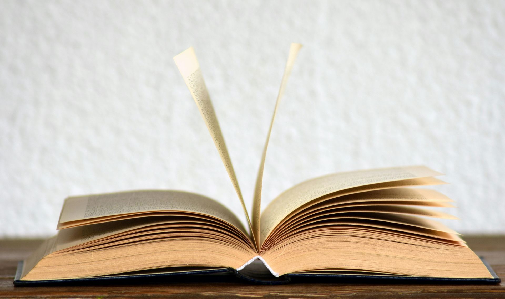 Open book on a wooden surface, pages slightly fanned, against a white wall.