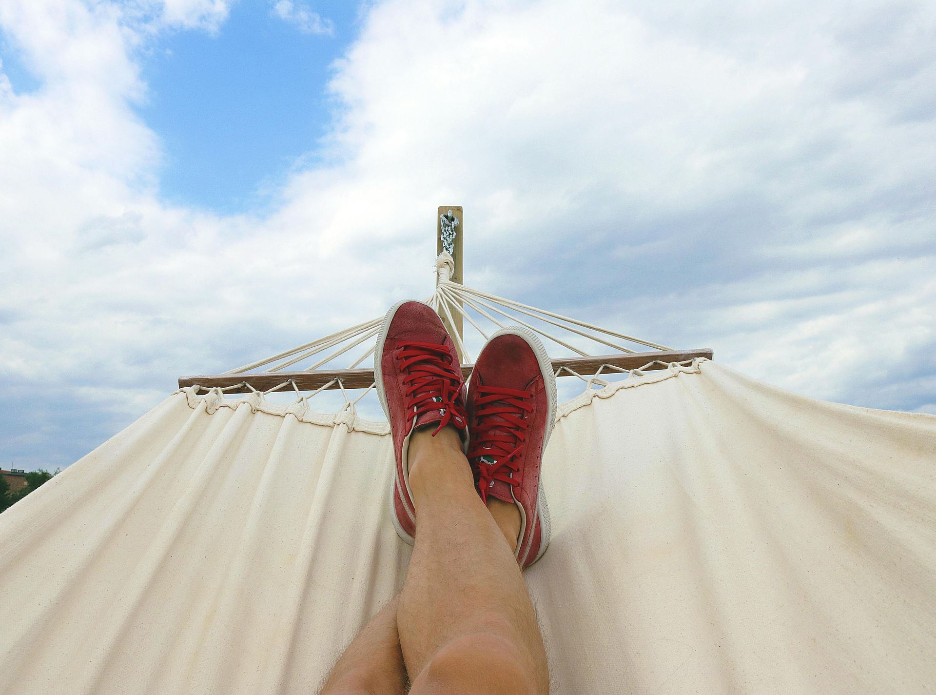 Person relaxing in a hammock, red sneakers, blue sky with clouds.