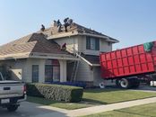 Workers on the roof of a house under renovation with a large red construction dumpster parked in the front yard.