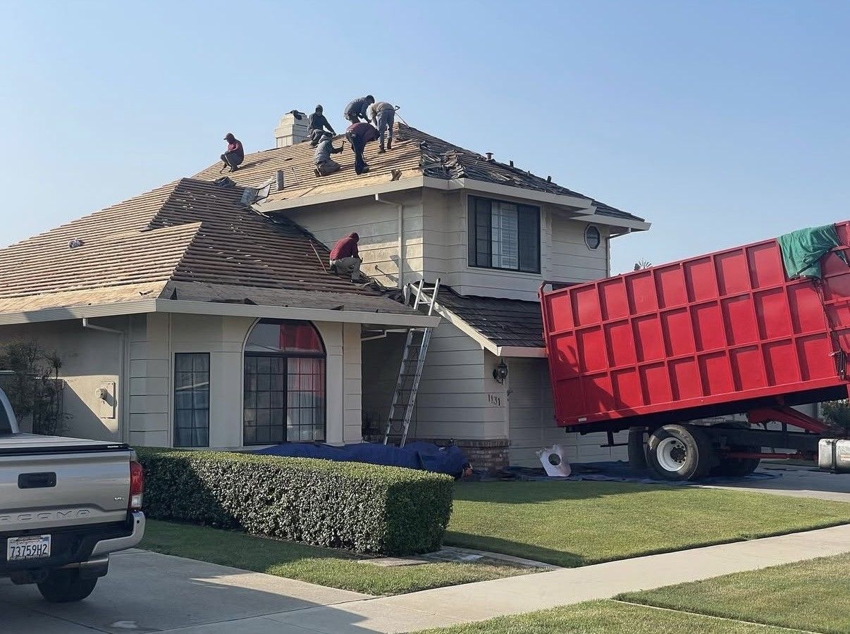 Workers on the roof of a house under renovation with a large red construction dumpster parked in the front yard.