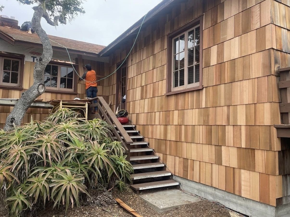 A worker in an orange vest installs wood shingles on the exterior wall of a building next to an outdoor wooden staircase.