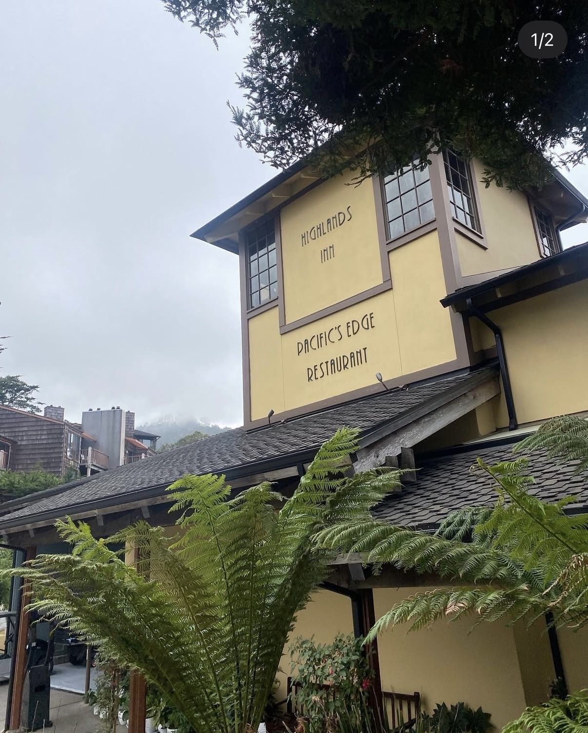 A pale yellow, multi-story building with dark tiled roofs and an exterior sign, surrounded by lush green plants.