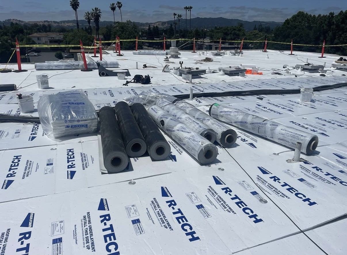 Construction materials, including R-Tech insulation panels and rolls, sit on a white flat roof under a clear blue sky.