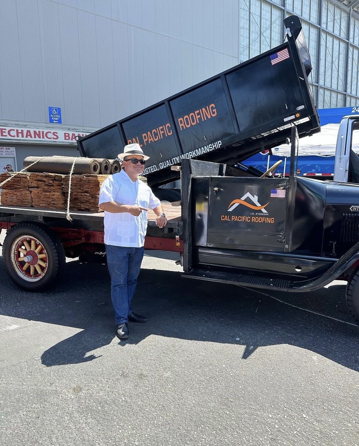 A person in a hat and white shirt stands next to a classic black dump truck with an orange logo in a parking lot.