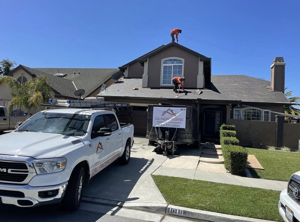 Workers repair the roof of a suburban house with a company truck and trailer parked in the driveway.