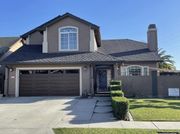 A two-story tan suburban house with a dark brown garage door, tiled roof, and manicured green hedges along the walkway.