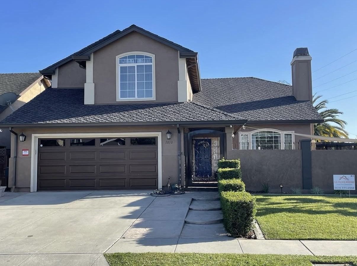A two-story tan suburban house with a dark brown garage door, tiled roof, and manicured green hedges along the walkway.