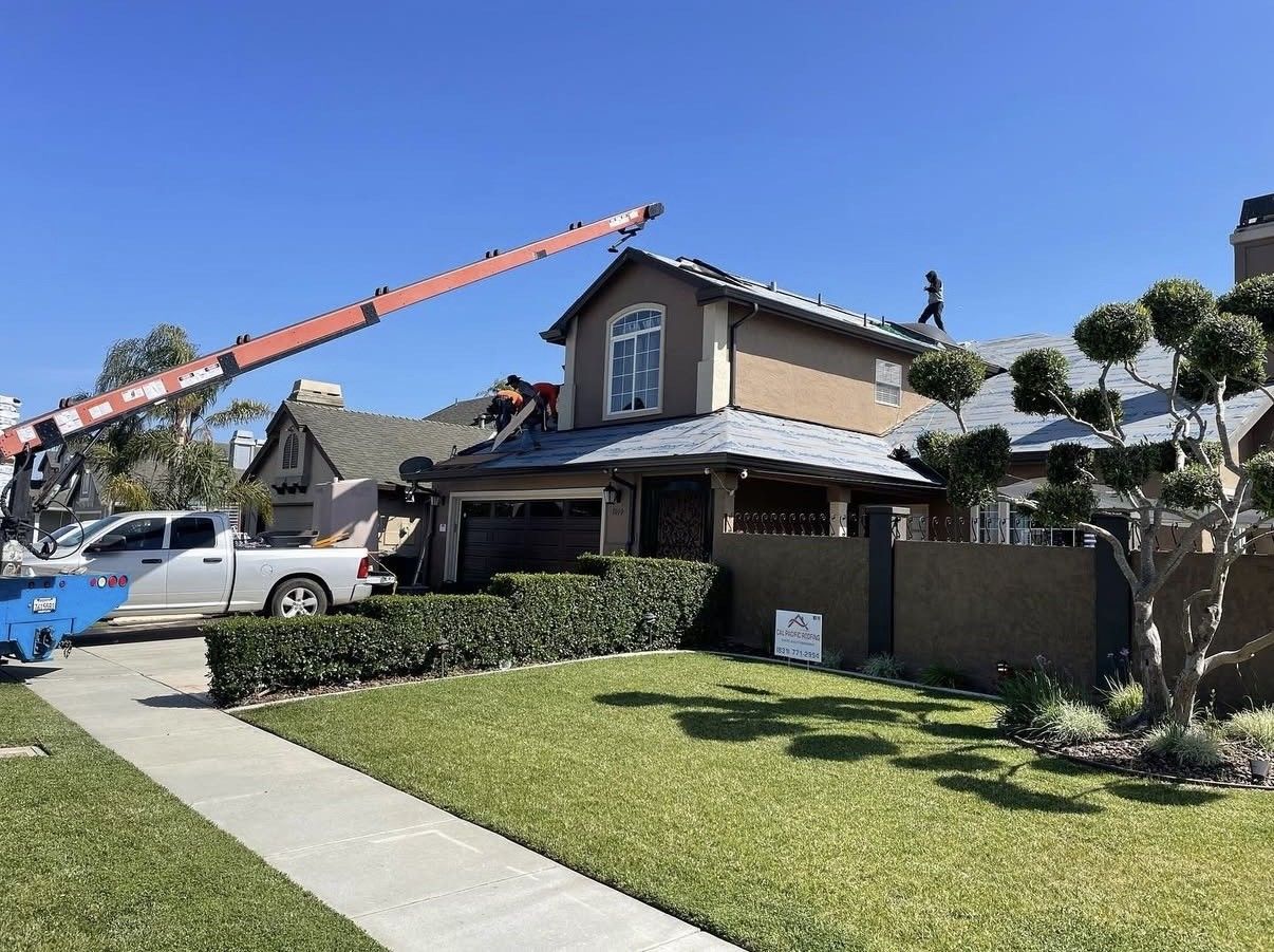 A white truck with a large crane performs roof repairs on a two-story house under a clear blue sky.