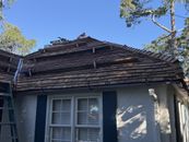 A ladder stands against the side of a stucco house with a wood shingle roof under a clear blue sky.