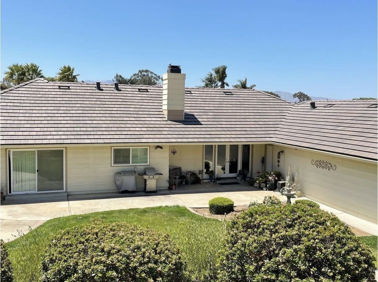 A beige, single-story house with a brown tiled roof, a large central chimney, and a concrete patio in the backyard.