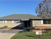 A tan, single-story suburban house with a gray shingled roof, a paved driveway, and a real estate sign on the front lawn.