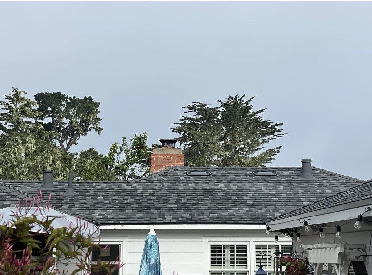 A white house with a gray shingle roof and a red brick chimney, set against a backdrop of green trees under a cloudy sky.