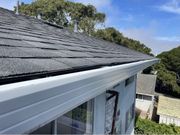 A low-angle view of a grey shingled roof with a white gutter running along the edge against a blue sky and trees.