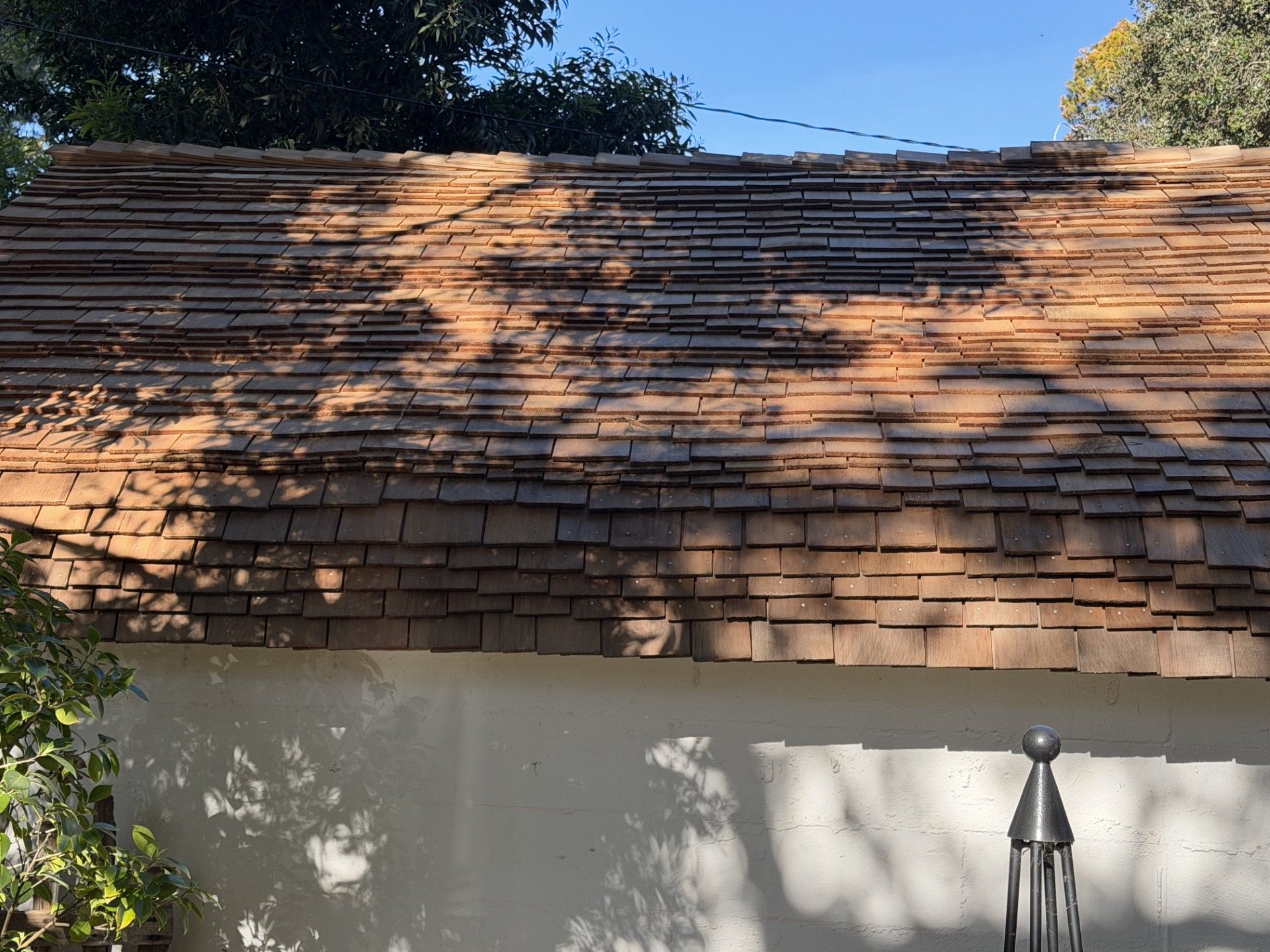 A weathered, layered wood shake roof on a building with a white stucco wall, partially shaded by tree leaves.
