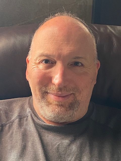 Bald man with a slight beard smiles, looking forward. He wears a gray shirt, against a brown background.
