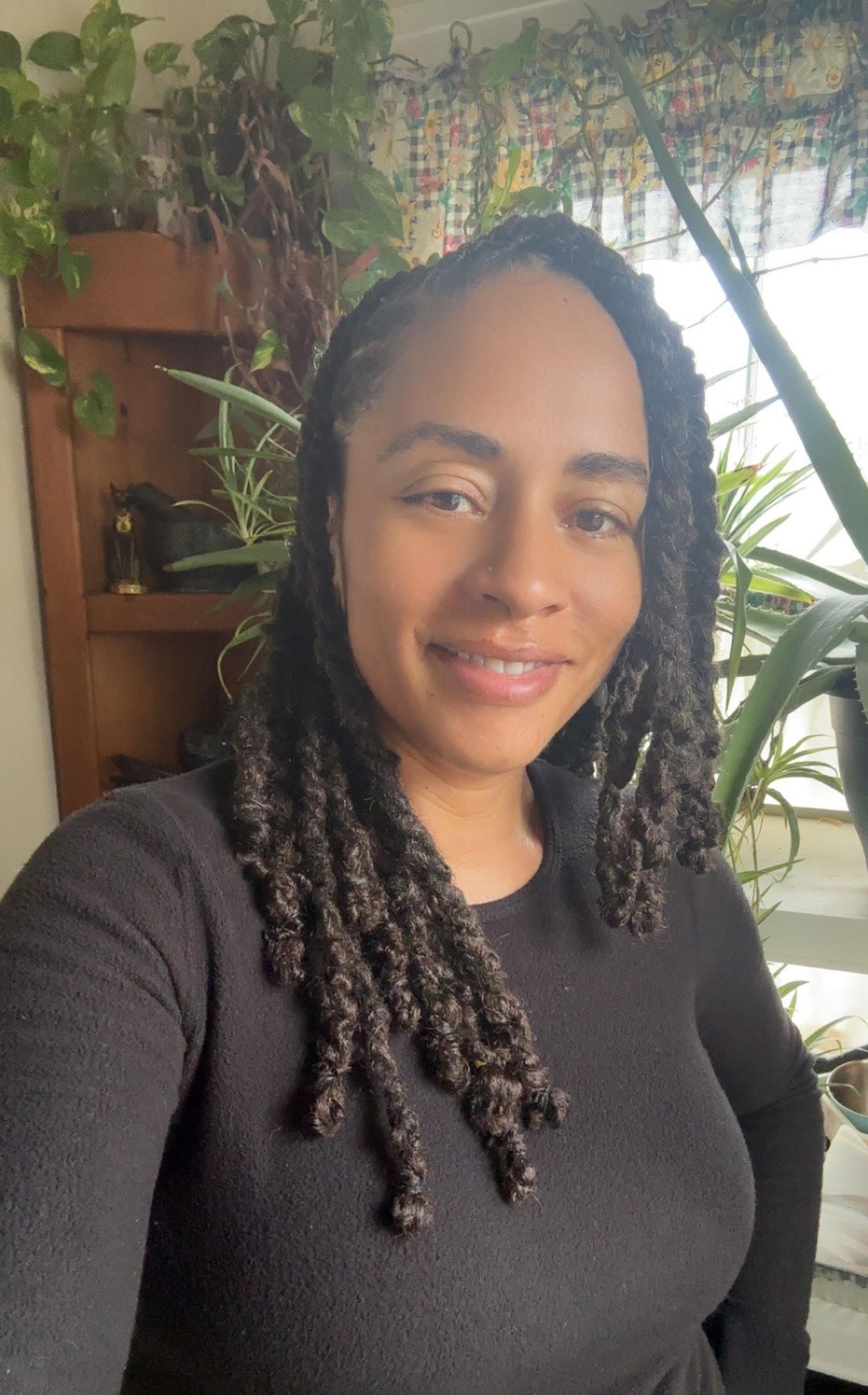 Woman with long braids in a black sweater smiles. Indoor setting with plants and a wooden shelf.
