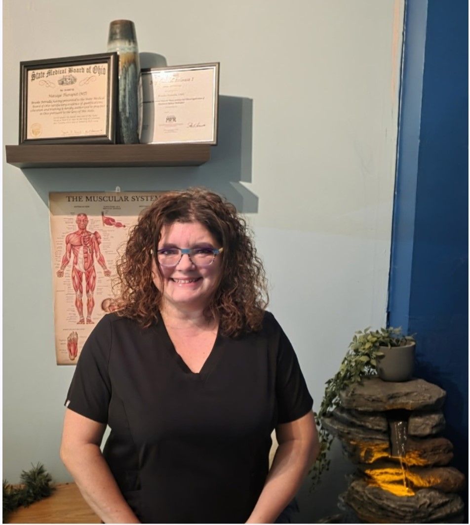 Woman in scrubs smiles in a room with medical chart and certificates on the wall.