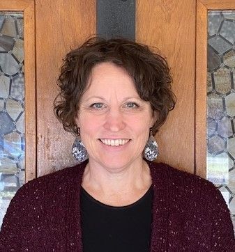 Woman with curly brown hair smiles, wearing dark sweater, and patterned earrings, standing in front of a wood door.