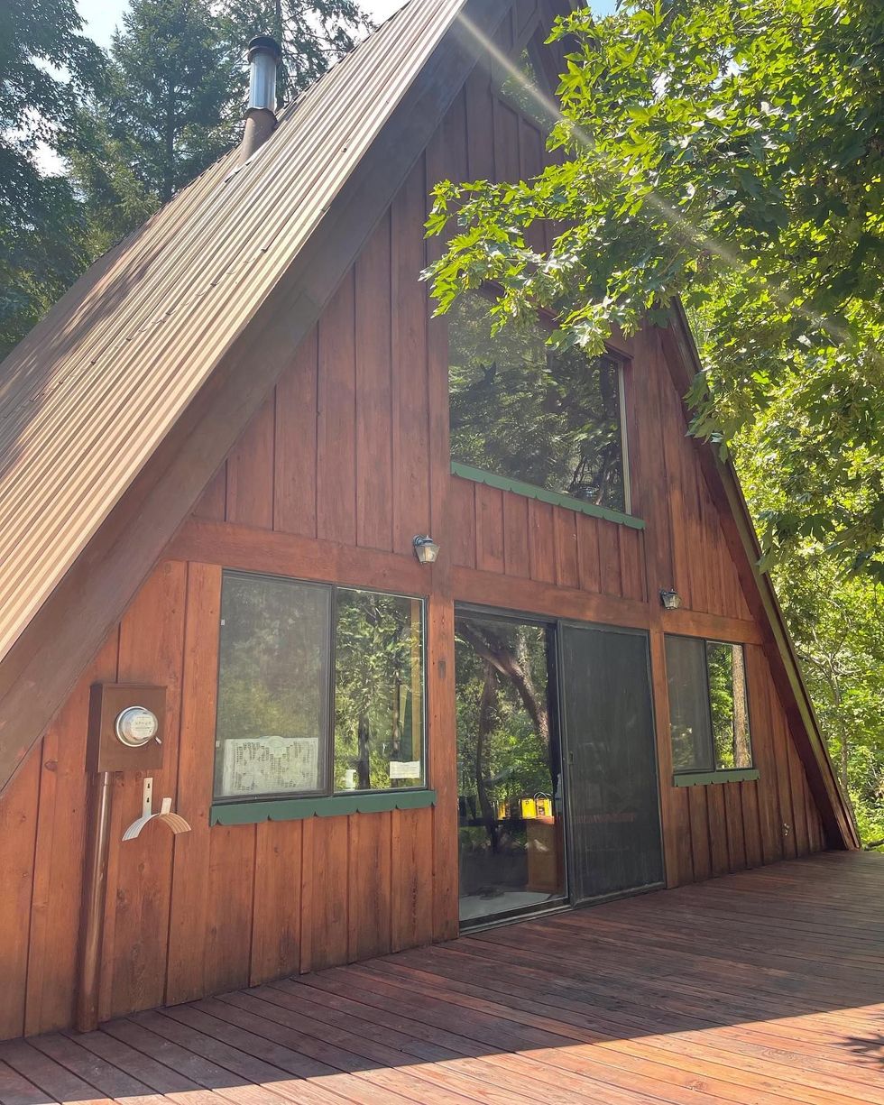 A-frame cabin with a wooden exterior and deck, tall windows, and a surrounding forest.