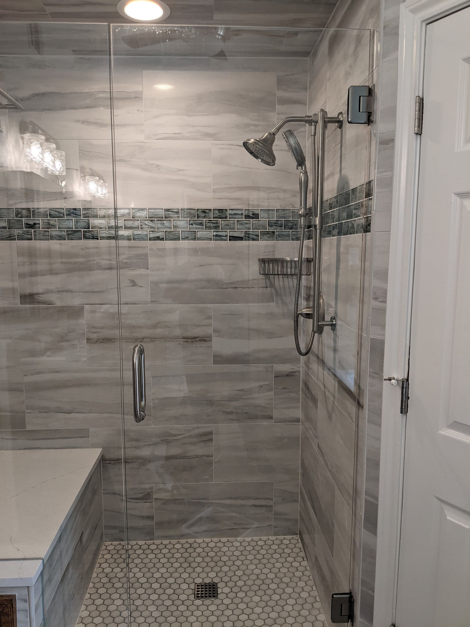 Shower with gray tiled walls, glass door, bench, and silver shower fixtures.