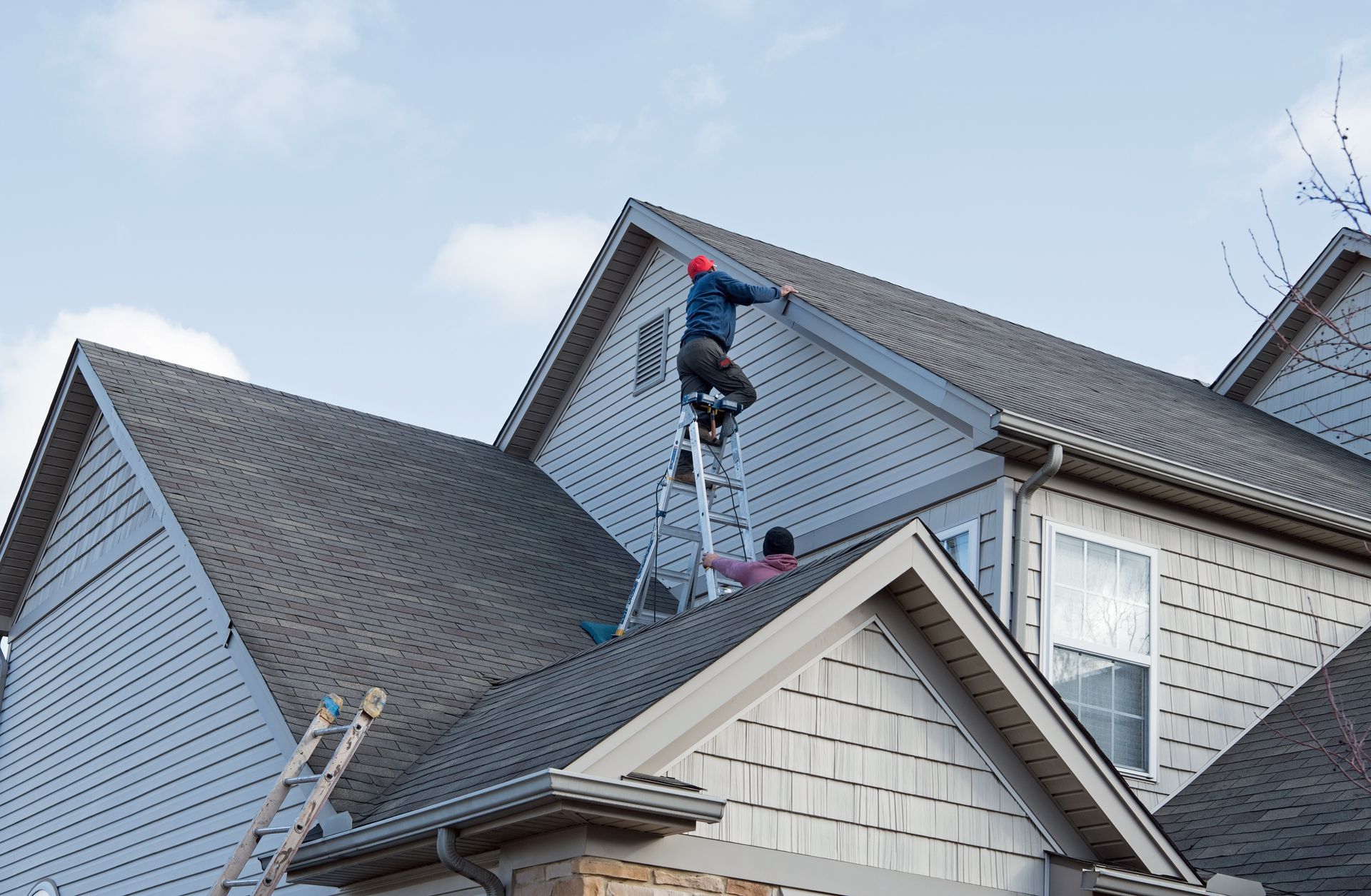 Two people on ladders working on the roof of a house, under a blue sky.