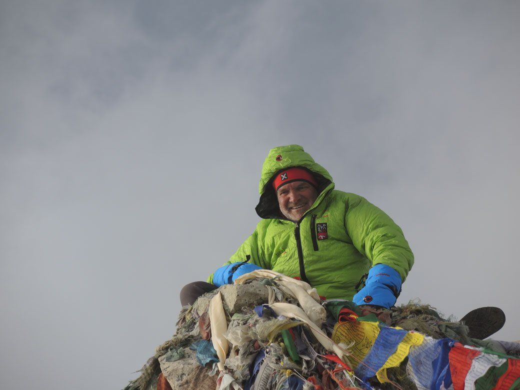 Ein Mann in einer grünen Jacke sitzt auf einem Berggipfel.