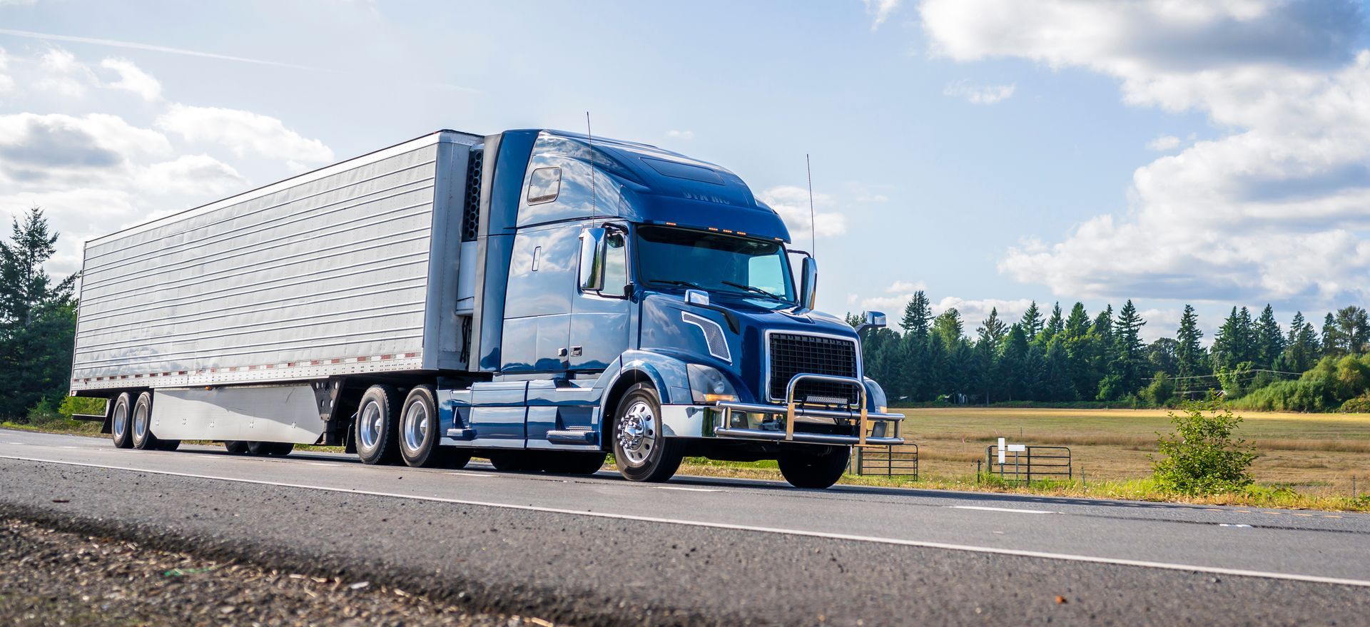 Blue semi-truck driving on a road with trees and fields in the background. Sunny day.