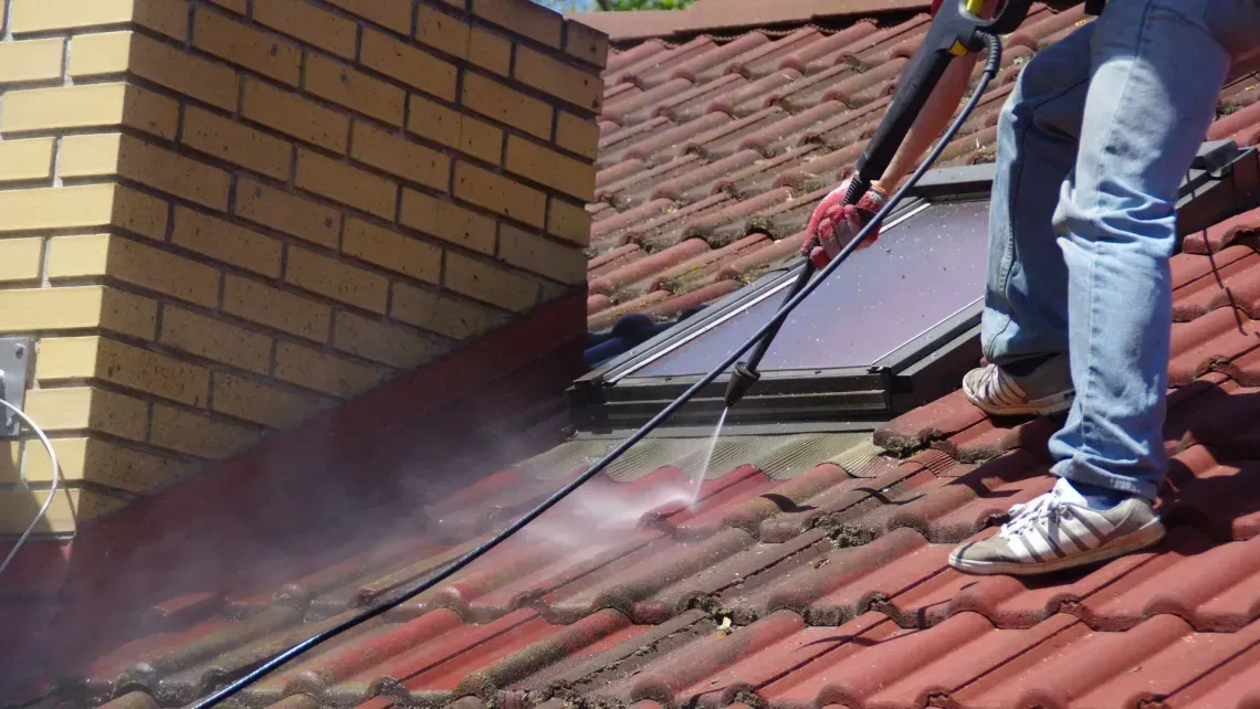 Person pressure-washing a red tiled roof beside a brick chimney.