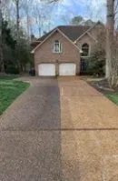 Brick suburban house with two-car garage and a long gravel driveway bordered by trees and winter grass