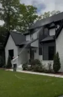Person spraying a two-story gray house with a hose in a green front yard.