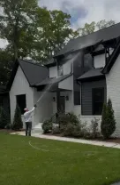 Person spraying a two-story gray house with a hose in a green front yard.