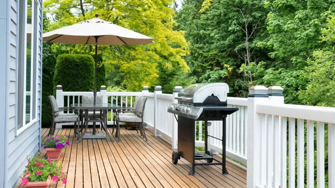 Wooden deck with patio table, umbrella, grill, and white railing beside green trees