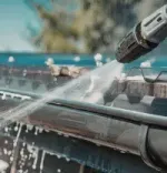 Pressure washing a roof gutter with a spray nozzle, water splashing over dark shingles