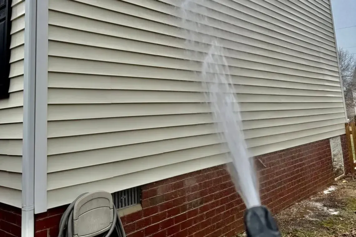 Person spraying water on beige house siding above red brick foundation with a hose nozzle