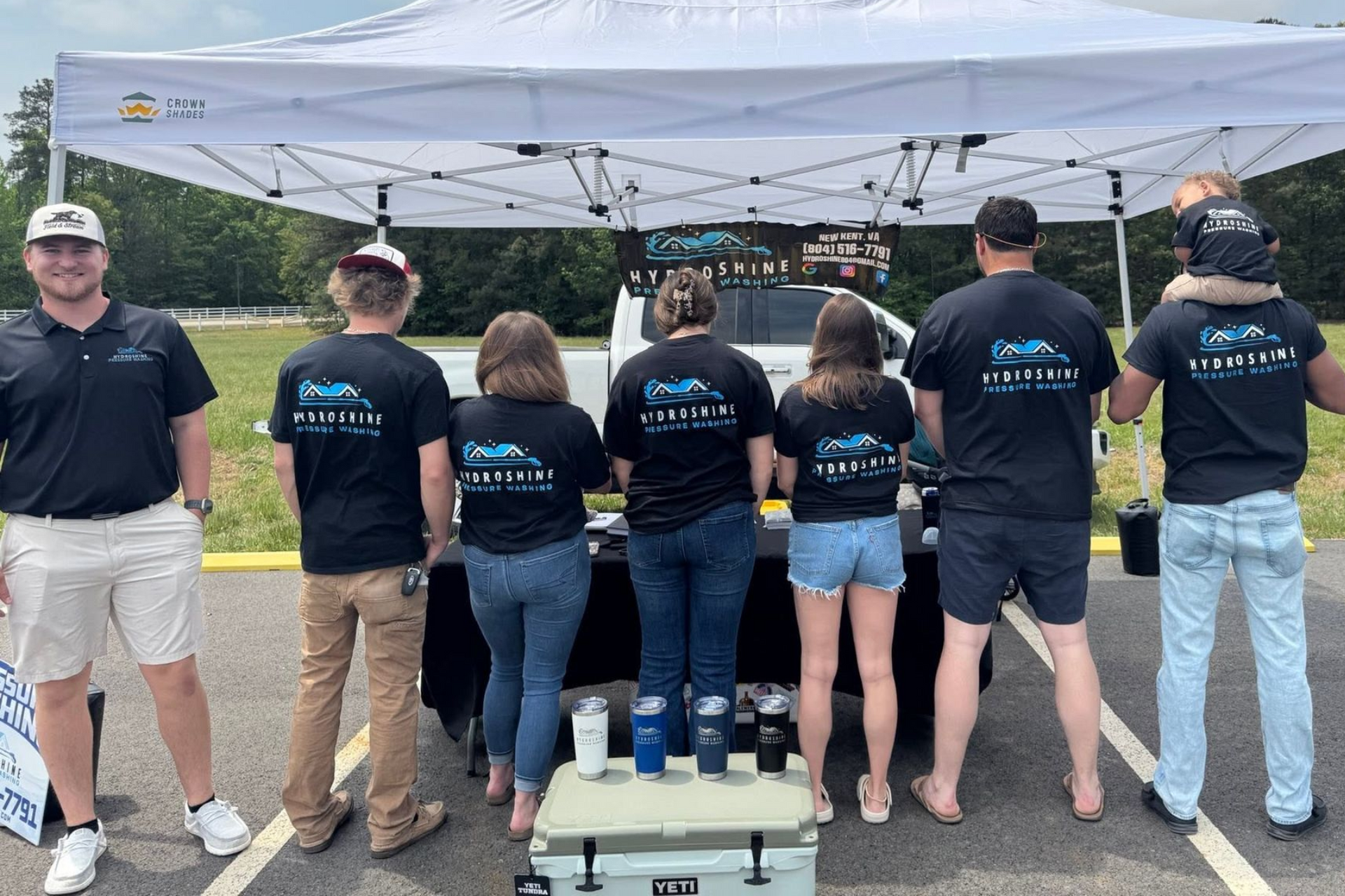 Volunteers in black shirts under a white tent at an outdoor event booth, seen from behind.