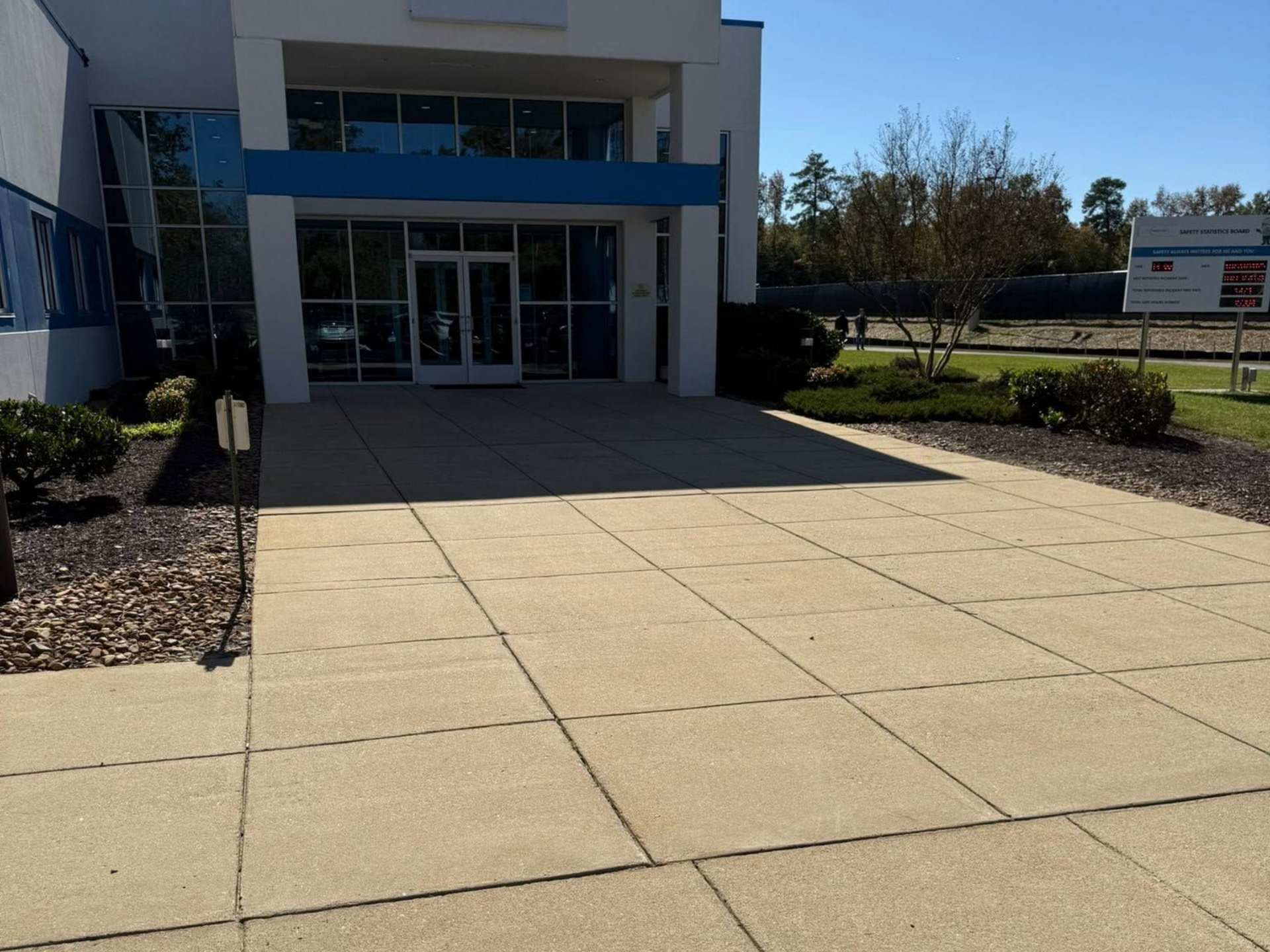 Modern building entrance with glass doors, blue awning, and a wide concrete walkway bordered by mulch beds