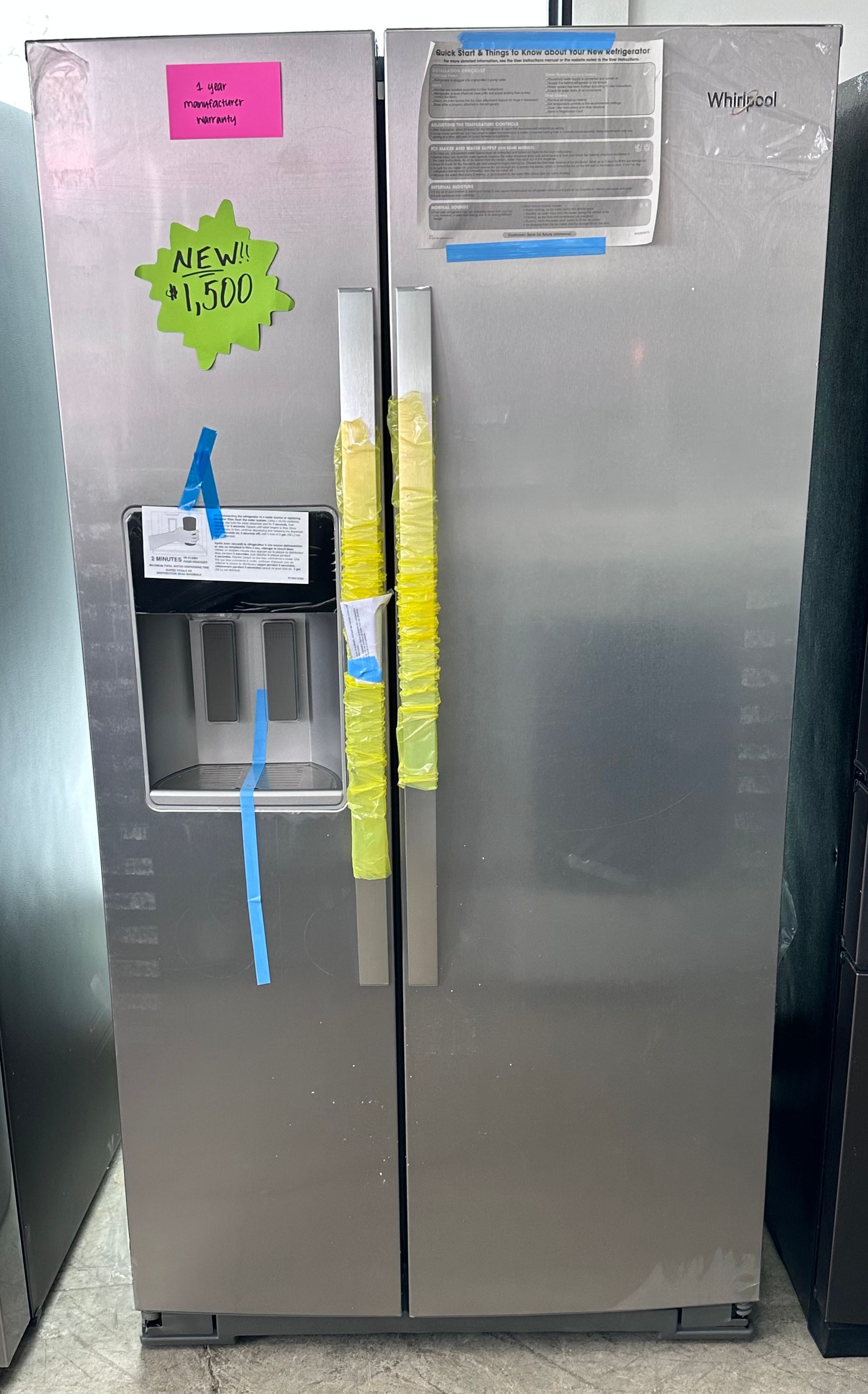 Side-by-side Whirlpool refrigerator. Stainless steel doors, ice/water dispenser, price tags, and yellow tape on handles.