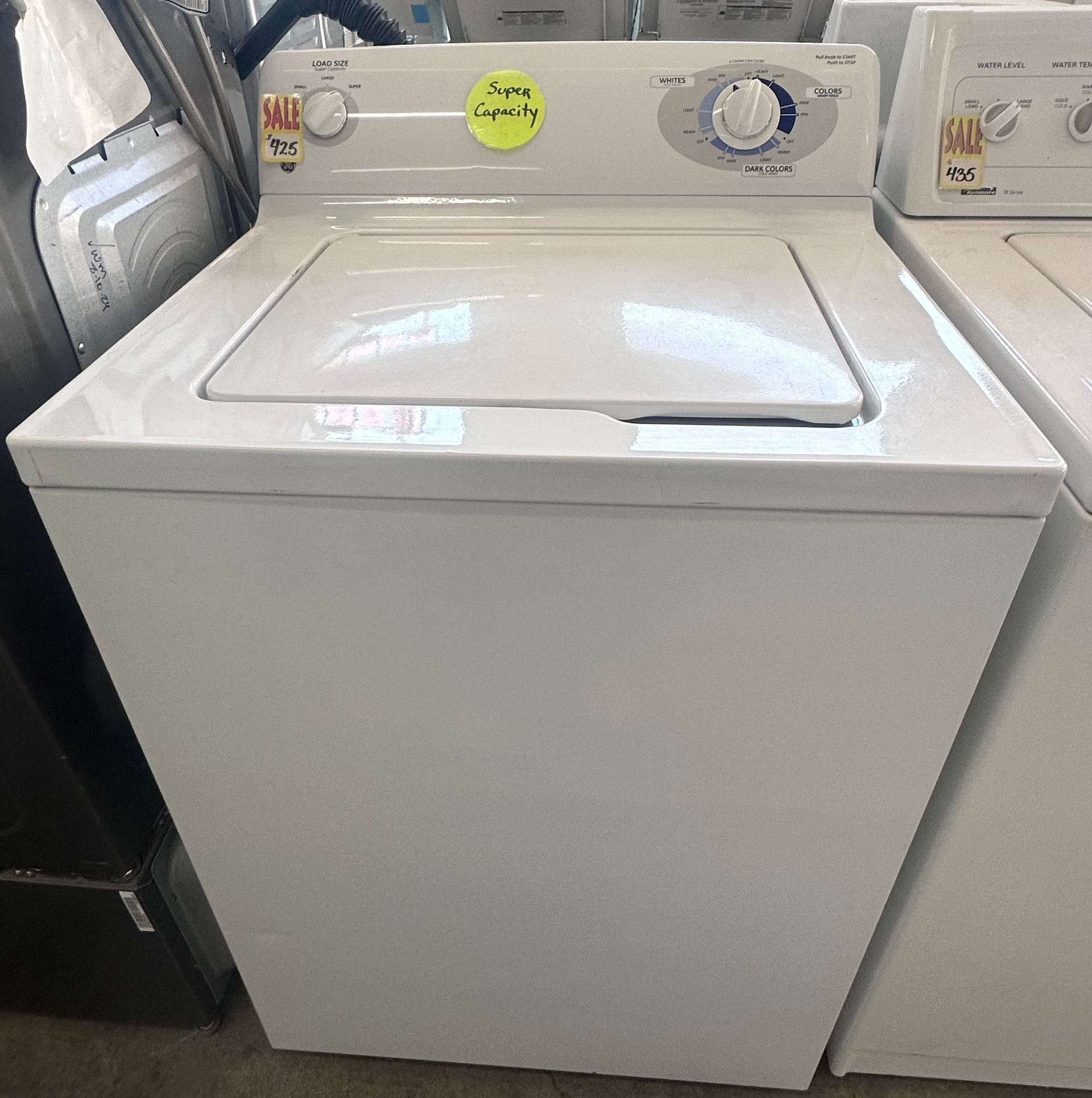 White top-load washing machine with control panel and lid, shown in a showroom.