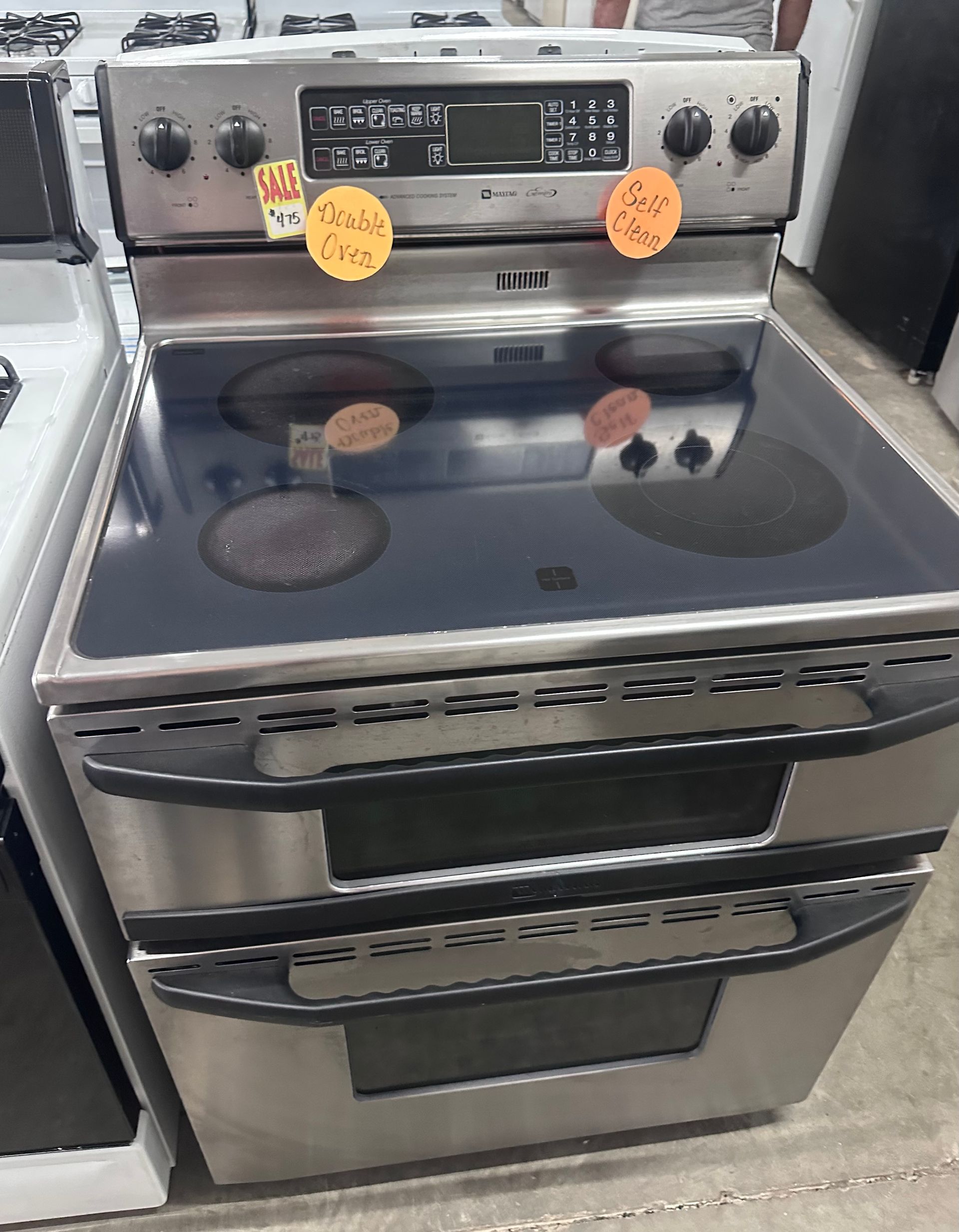 Stainless steel electric range with glass cooktop and oven, shown in a store aisle with sale tags.