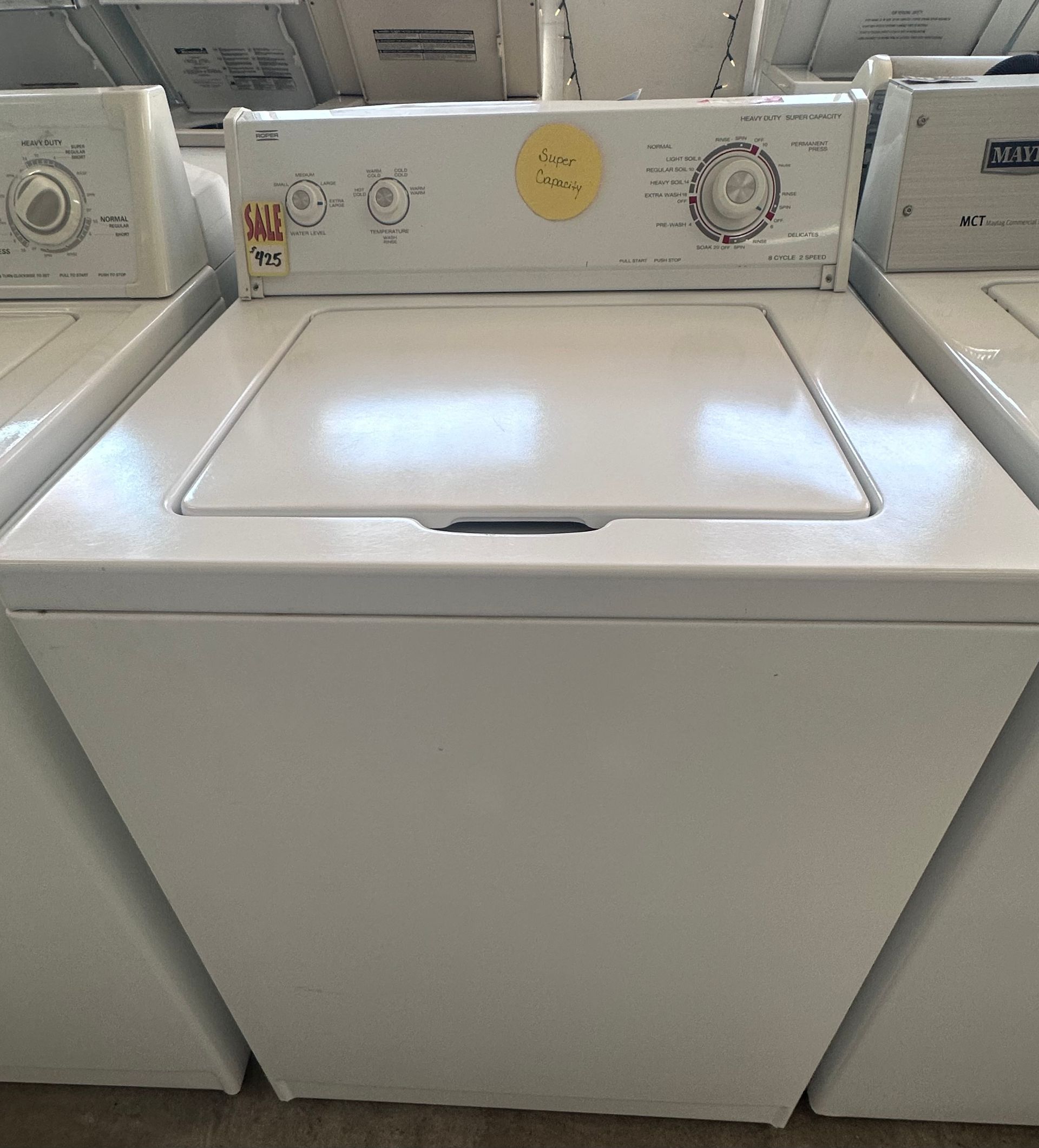 White top-loading washing machine in a laundromat aisle, with control panel and lid visible