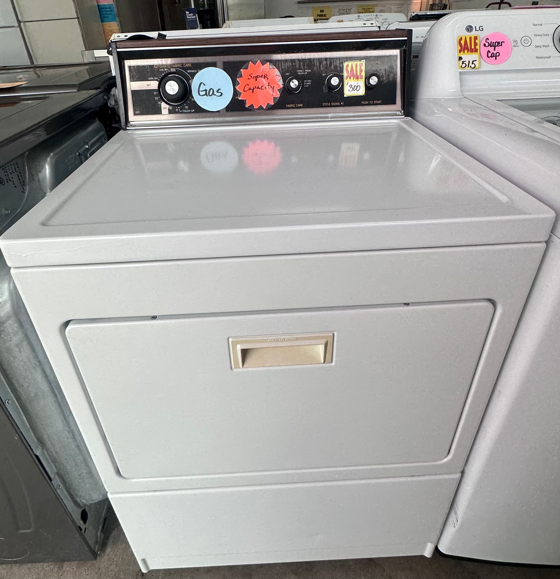 White top-load dryer in an appliance store, with control panel and drawer handle visible.